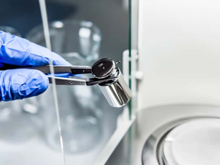 Kalibrierung Prüfgewichte 2 Die Hand eines Bedieners hält ein Stahlkalibriergewicht, um es auf die Analysenwaage zu legen.