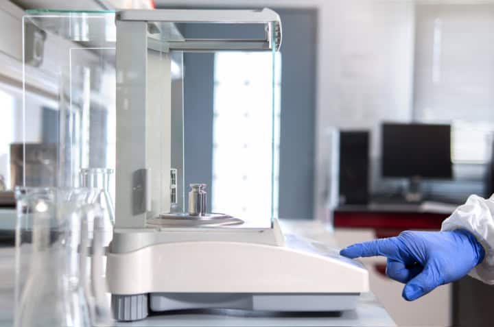 Waagen Kalibrierung 1 Die Hand eines Bedieners hält ein Stahl-Kalibriergewicht, um es auf die Analysenwaage zu legen.