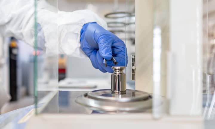 Start 1 Eine Hand eines Bedieners hält ein Stahl-Prüfgewicht, um es auf die Analysenwaage zu legen.